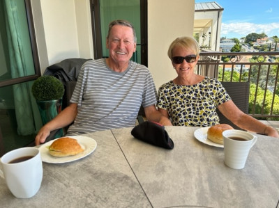 A man in a striped shirt (Mark) and a woman wearing a leaf-print top (Beth) and sunglasses sitting at an outdoor table on a deck smiling over coffee.